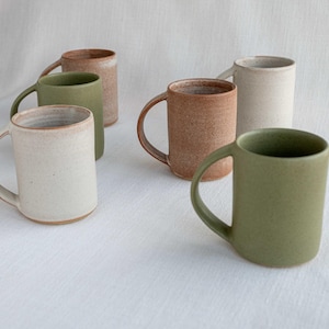 May include: Six ceramic mugs in various colors, including green, brown, and white. The mugs are all similar in size and shape, with a rounded body and a handle.