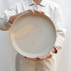 May include: A large, round, speckled ceramic plate with a matte, off-white finish. The surface features a smooth, lighter-colored, curved abstract glaze pattern that sweeps across the left side of the plate, contrasting with the textured base.