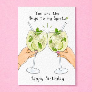 May include: A greetings card featuring a watercolour illustration of two hands holding glasses filled with a light yellow cocktail, lime slices, mint leaves, and straws. The text reads, "You are the Hugo to my Spritz" and "Happy Birthday."