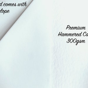 以下が含まれることがあります： 「Card comes with Envelope」と「Premium Hammered Card 300gsm」の文字が入った白いカードのクローズアップ。カードはテクスチャ加工された表面で、画像では斜めに配置されています。