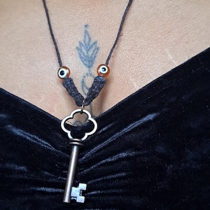 Könnte beinhalten: Eine schwarze Choker-Kette mit einem silbernen Schlüsselanhänger und kleinen Perlen. Die Kette besteht aus einem dünnen schwarzen Band und hat einen silbernen Schlüsselanhänger mit einem dekorativen Design. Der Schlüssel ist mit einem kleinen silbernen Ring am Band befestigt. Die Kette hat auch kleine Perlen in verschiedenen Farben, darunter Schwarz, Weiß und Grün.