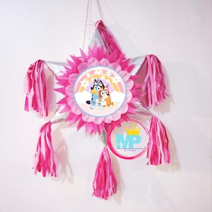 May include: A pink and silver star-shaped piñata with a colorful cartoon image of two dogs. The piñata has pink and white tassels hanging from it. The text "M Piñata" is printed on the bottom of the piñata.