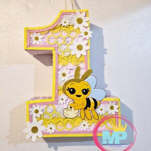 May include: A large number one pi&ntilde;ata, decorated with a bee and flowers. The pi&ntilde;ata is yellow, pink, and white, with a honeycomb pattern. A cartoon bee holds a basket. The pi&ntilde;ata is designed for a first birthday celebration.