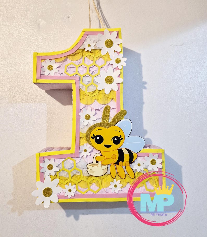 May include: A large number one pi&ntilde;ata, decorated with a bee and flowers. The pi&ntilde;ata is yellow, pink, and white, with a honeycomb pattern. A cartoon bee holds a basket. The pi&ntilde;ata is designed for a first birthday celebration.