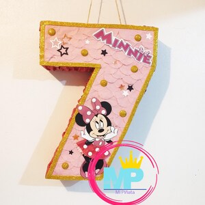 Puede incluir: Una pi&ntilde;ata rosa con el n&uacute;mero siete, con adornos dorados y gr&aacute;ficos de Minnie Mouse. La pi&ntilde;ata tiene una textura de vieira y est&aacute; decorada con estrellas y c&iacute;rculos dorados. La palabra "Minnie" se muestra en la parte superior.