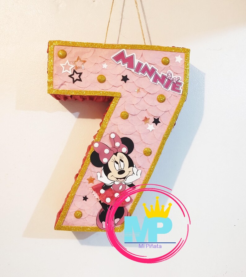 Puede incluir: Una pi&ntilde;ata rosa con el n&uacute;mero siete, con adornos dorados y gr&aacute;ficos de Minnie Mouse. La pi&ntilde;ata tiene una textura de vieira y est&aacute; decorada con estrellas y c&iacute;rculos dorados. La palabra "Minnie" se muestra en la parte superior.