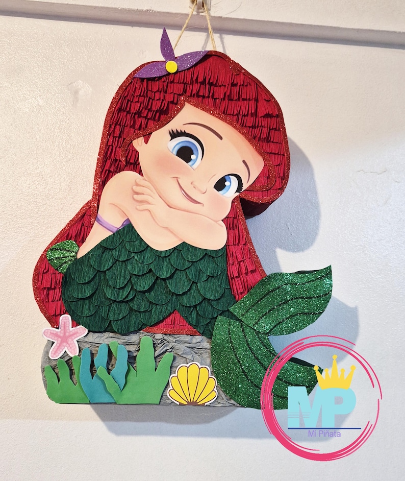 May include: A handcrafted pi&ntilde;ata shaped like a cartoon mermaid with bright red hair, a green scale-textured tail, and a purple flower accessory. The base features paper cutouts of a pink starfish, green seaweed, and a yellow seashell.
