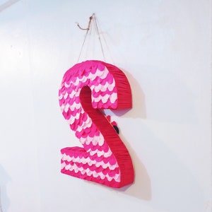 Puede incluir: Una pi&ntilde;ata rosa grande con forma del n&uacute;mero dos, decorada con escamas de papel rosas y blancas alternas. La pi&ntilde;ata est&aacute; colgada de un gancho en una pared blanca, ideal para una fiesta de cumplea&ntilde;os.
