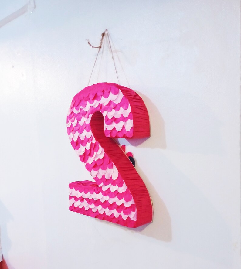 Puede incluir: Una pi&ntilde;ata rosa grande con forma del n&uacute;mero dos, decorada con escamas de papel rosas y blancas alternas. La pi&ntilde;ata est&aacute; colgada de un gancho en una pared blanca, ideal para una fiesta de cumplea&ntilde;os.