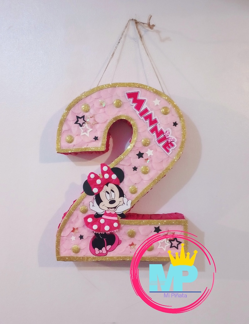 Puede incluir: Una pi&ntilde;ata rosa con el n&uacute;mero 2, borde dorado con purpurina y Minnie Mouse. La pi&ntilde;ata est&aacute; decorada con estrellas y c&iacute;rculos dorados. La palabra "Minnie" est&aacute; escrita en letras rojas. Un art&iacute;culo decorativo para una fiesta de cumplea&ntilde;os.