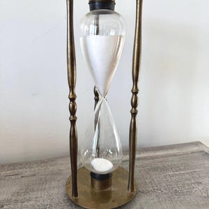 May include: An antique hourglass with a brass frame and clear glass bulbs. The hourglass is filled with white sand and is supported by four brass rods. The top and bottom are circular brass plates.