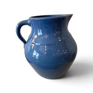 May include: A blue ceramic pitcher with a curved handle. The pitcher has a glossy finish and a rounded body. The pitcher is approximately 8 inches tall and 6 inches wide.