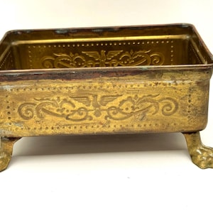 May include: A rectangular, antique brass container with ornate embossed designs and claw feet. The container has a gold finish with signs of wear, and decorative patterns including an eagle and floral motifs.