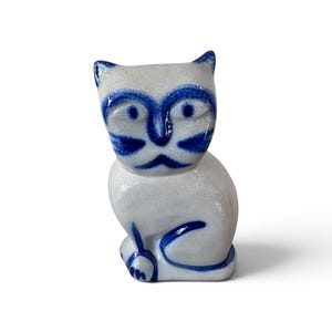 May include: A small ceramic cat figurine with a speckled off-white glaze and hand-painted cobalt blue details. The cat features large, wide-set eyes, a distinct nose, and whiskers, with a curled tail resting against its side in a seated position.