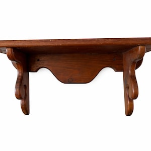 May include: A small, wooden shelf with a dark brown finish. The shelf has a rectangular top and decorative, curved supports. The supports are attached to the underside of the shelf and feature a scroll-like design. The shelf is designed for wall mounting.