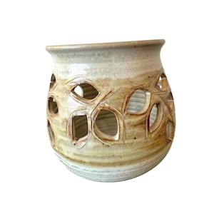 May include: A handcrafted ceramic vase with a light beige and brown color scheme. The vase features leaf-shaped cutouts around the body, creating a decorative pattern. The rim is smooth, and the overall shape is rounded.