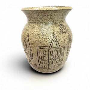 May include: A speckled beige ceramic vase with a wide mouth and a rounded body. The vase features etched drawings of a building, trees, and a rose. The vase is a decorative item.