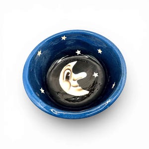 May include: A blue ceramic bowl with a whimsical design. The bowl features a black interior with a smiling crescent moon and white stars. The exterior is a solid blue color.