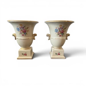 May include: Two cream-colored ceramic urns with floral designs in pink, blue, and purple. Each urn has gold trim and handles, and sits on a square base with a small floral accent. A classic decorative item.