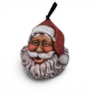 May include: A decorative Santa Claus ornament featuring a sculpted face with a long white beard, rosy cheeks, and a red hat with a white trim and pom-pom. The ornament includes a black ribbon loop at the top for hanging, showcasing detailed facial features.