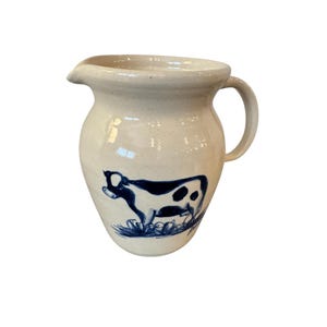 May include: Cream-colored ceramic pitcher with a blue cow illustration. The pitcher has a rounded body, a spout, and a curved handle. The cow is depicted in a simple, stylized manner, with black and white markings. The pitcher is suitable for serving beverages.
