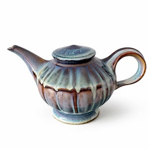 May include: A ceramic teapot with a lid, spout, and handle. The teapot has a ribbed design and is glazed in shades of blue, brown, and green. The handle is a loop shape. The teapot sits on a small, round base.