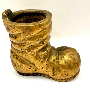 May include: A vintage brass boot-shaped container with a textured surface and a worn, aged appearance. The boot has a wide opening at the top and is likely used for decorative purposes. The brass has a golden hue with areas of green oxidation.