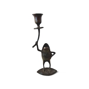 May include: A bronze-colored frog-shaped candlestick holder. The frog stands on a leaf-shaped base, holding a candle cup aloft. The candlestick is made of metal and has a vintage appearance.