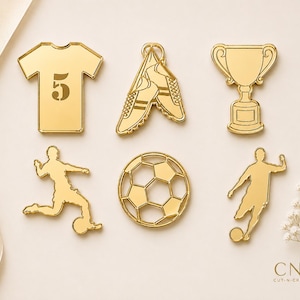 以下が含まれることがあります： 背番号5のジャージ、スパイク、トロフィー、サッカーボール、ボールを蹴る選手のシルエット2体というサッカーをテーマにした6つのゴールドカラーのアクリルチャームセット。CNC CUT-N-CRAFT.UKの文字が記されています。