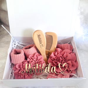 May include: A white gift box containing baby items. Inside are pink socks, two wooden hairbrushes, and two large flower hair accessories. A gold nameplate with the name "Matilda" is also included.