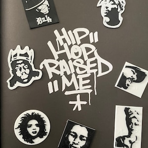 May include: A collection of black and white portraits of famous hip hop artists, including Tupac Shakur, Biggie Smalls, and others. The portraits are surrounded by the words "Hip Hop Raised Me" in white spray paint.