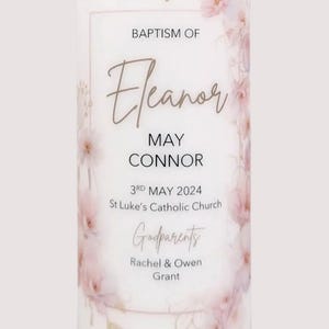 May include: A white baptism candle featuring a gold cross and delicate pink floral decorations. The candle is inscribed with the name "Eleanor", date, church details, and godparents' names. A pink ribbon adorns the base.