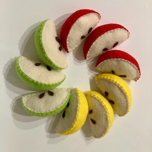 May include: A circular arrangement of eight felt apple slices in red, green, and yellow. Each slice features a cream-colored interior with visible brown felt seeds and stitched edges, creating a soft, textured appearance for each fruit piece.