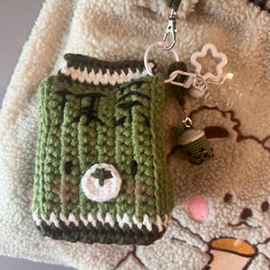 May include: A green crochet case with a bear face design, white and dark green trim, and a silver clasp. The case is attached to a white star-shaped charm and a small green crochet figure. The background is a cream-colored, textured fabric.