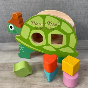 May include: A green wooden turtle shape sorter toy with the name "Marnie Rose's Shape Sorter" engraved on the side. The toy comes with five colourful wooden shapes: a heart, a square, a triangle, a leaf, and a circle.