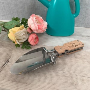 May include: A wooden folding gardening tool with a stainless steel trowel and a handle that says "The Chief Grower!" The tool is on a wooden surface with a teal watering can and pink flowers in the background.