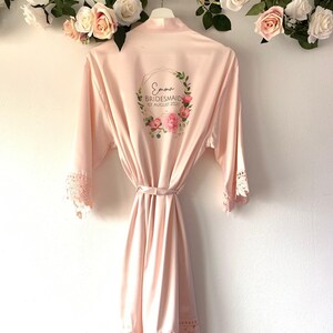 May include: A pale pink satin robe with lace trim and a floral wreath design on the back. The wreath includes the text "Emma Bridesmaid 1st August 2020".