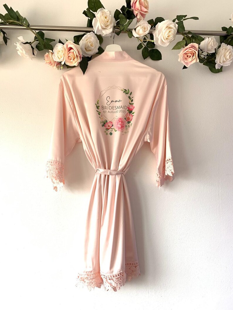 May include: A pale pink satin robe with lace trim and a floral wreath design on the back. The wreath includes the text "Emma Bridesmaid 1st August 2020".