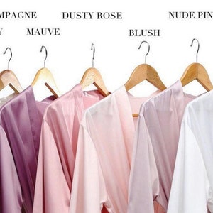 May include: Eight satin robes hanging on wooden hangers, displayed in a variety of colors, including light grey, mauve, dusty rose, blush, nude pink, and ivory. The robes are all made of a soft, silky fabric and have a classic kimono style.