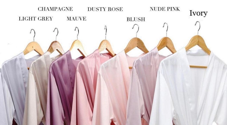 May include: Eight satin robes hanging on wooden hangers, displayed in a variety of colors, including light grey, mauve, dusty rose, blush, nude pink, and ivory. The robes are all made of a soft, silky fabric and have a classic kimono style.