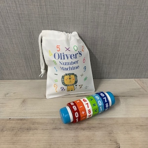 May include: A colourful wooden toy number machine with a lion illustration on the bag. The toy has a blue base and colourful rings with numbers and maths symbols. The bag is white with the text "Oliver's Number Machine" printed on it.