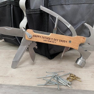 May include: A multi-tool with a wooden handle, featuring a knife, saw, hammer, and other tools. The handle is engraved with "HAPPY FATHER'S DAY DADDY LOVE FROM EVIE.X". Nails and screws are scattered nearby, with a black tool bag in the background.