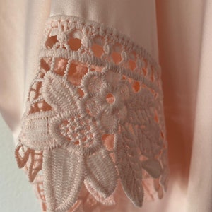 May include: Close-up of a pale pink satin fabric with a delicate lace trim. The lace is a floral design with a scalloped edge.