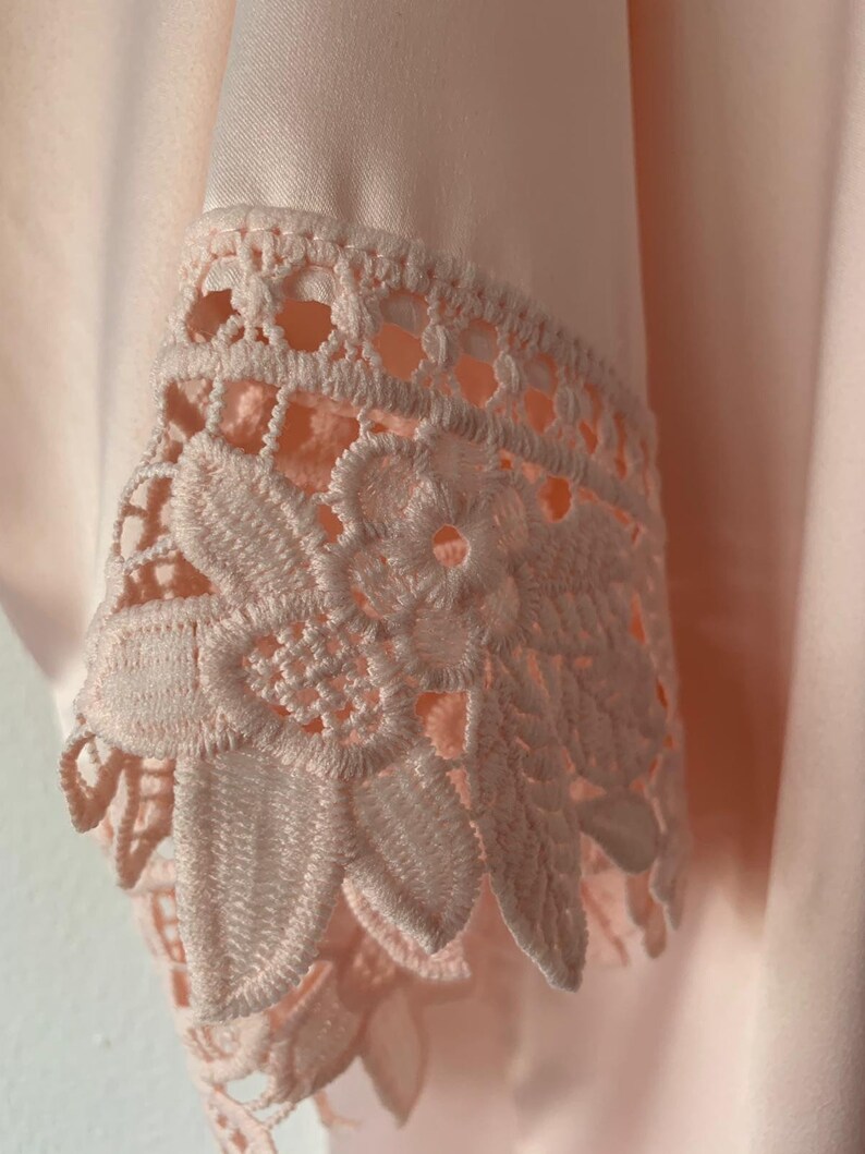 May include: Close-up of a pale pink satin fabric with a delicate lace trim. The lace is a floral design with a scalloped edge.