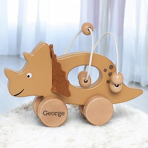 May include: A wooden toy triceratops with brown and tan paint, rolling wheels, and a string maze with wooden beads. The toy is personalised with the name "George" on one of the wheels.