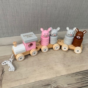 Puede incluir: Un tren de juguete de madera con bloques apilables de conejo rosa, gris y marrón. La locomotora tiene un techo blanco y un letrero que dice "Addison's Train".
