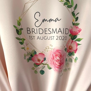 May include: A pink satin robe with a gold geometric frame and floral wreath design. The text "Emma Bridesmaid 1st August 2020" is printed on the robe.