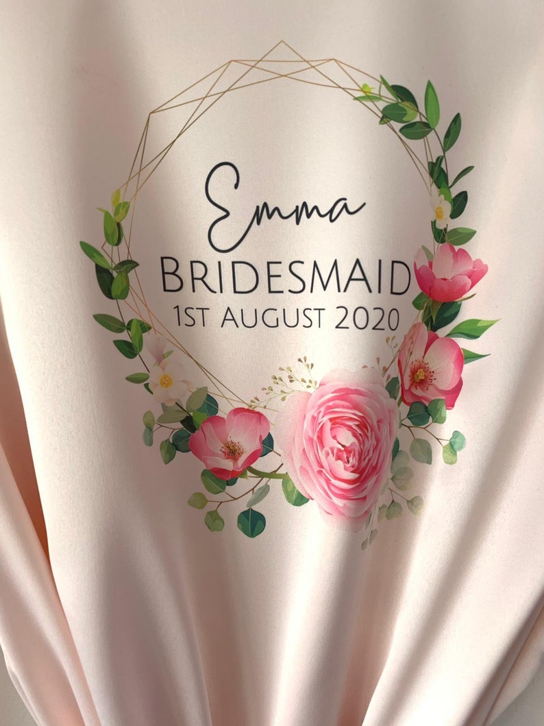 May include: A pink satin robe with a gold geometric frame and floral wreath design. The text "Emma Bridesmaid 1st August 2020" is printed on the robe.