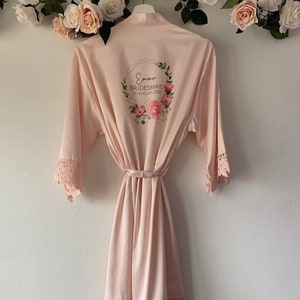 May include: A pale pink satin robe with lace trim and a floral wreath design on the back. The wreath includes the text "Emma Bridesmaid 1st August 2020".