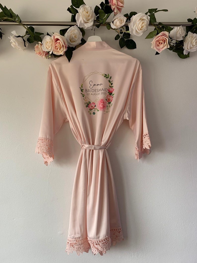 May include: A pale pink satin robe with lace trim and a floral wreath design on the back. The wreath includes the text "Emma Bridesmaid 1st August 2020".